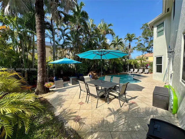 a view of patio with chairs and table under an umbrella with barbeque grill and large trees