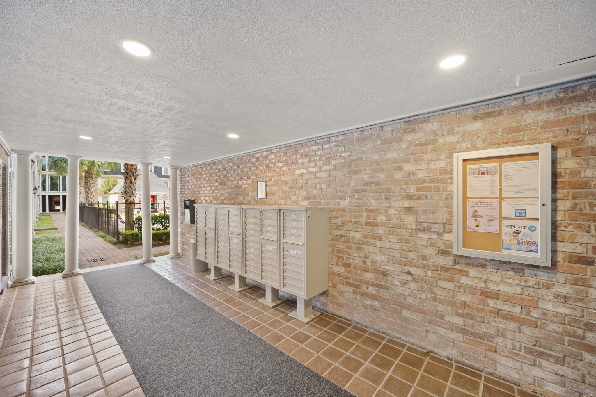 357 North Post Oak Lane, Unit 101 Houston, TX 77024 - Photo 13 of 19 A functional, covered breezeway area complete with secure grey cluster mailboxes against a light brick wall, an outdoor bulletin board, and modern recessed lighting.