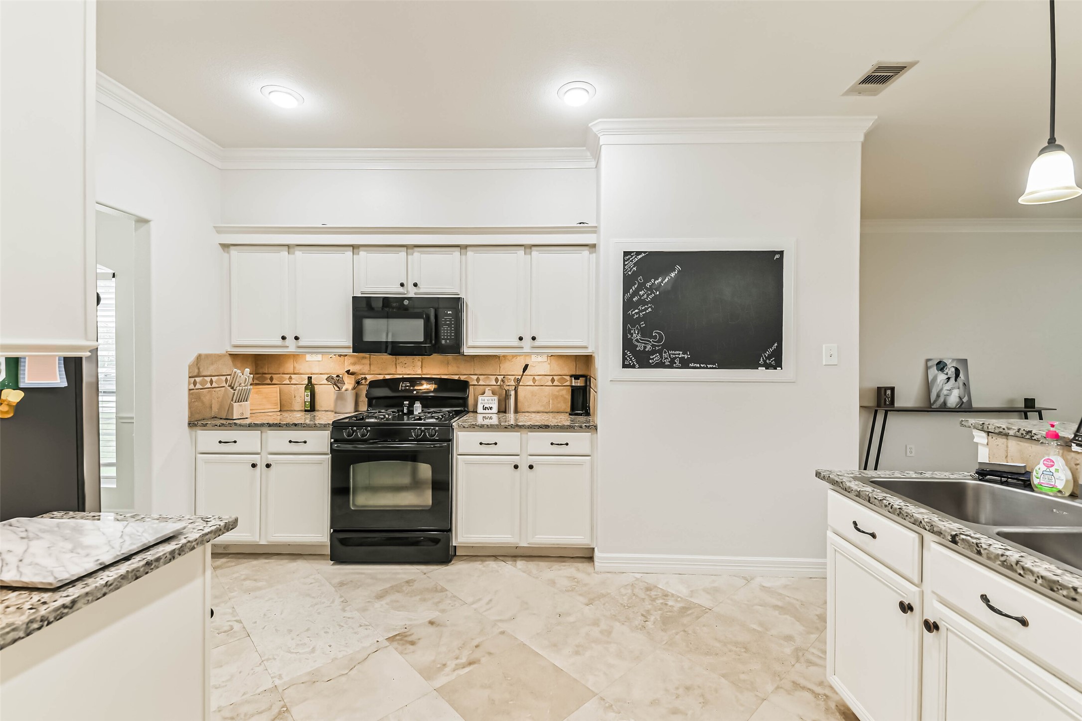 This kitchen features sleek white cabinetry, modern black appliances, and granite countertops. It includes a convenient chalkboard wall for notes and a cozy open layout, ideal for family gatherings.