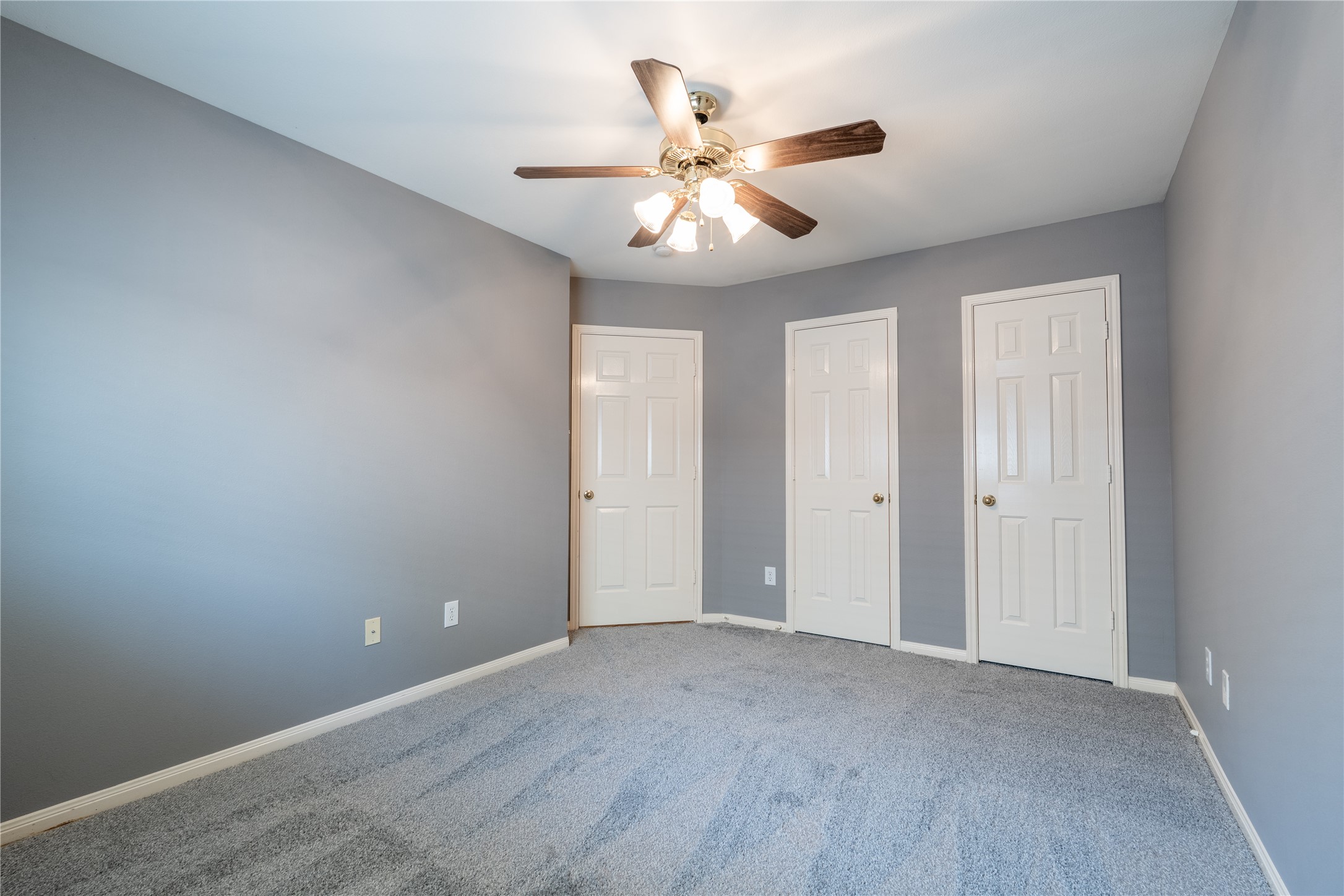 10619 Crownsedge Drive Spring, TX 77379 - Photo 24 of 38 This photo showcases a bright, empty room with gray walls and carpet, featuring a ceiling fan and three white doors.