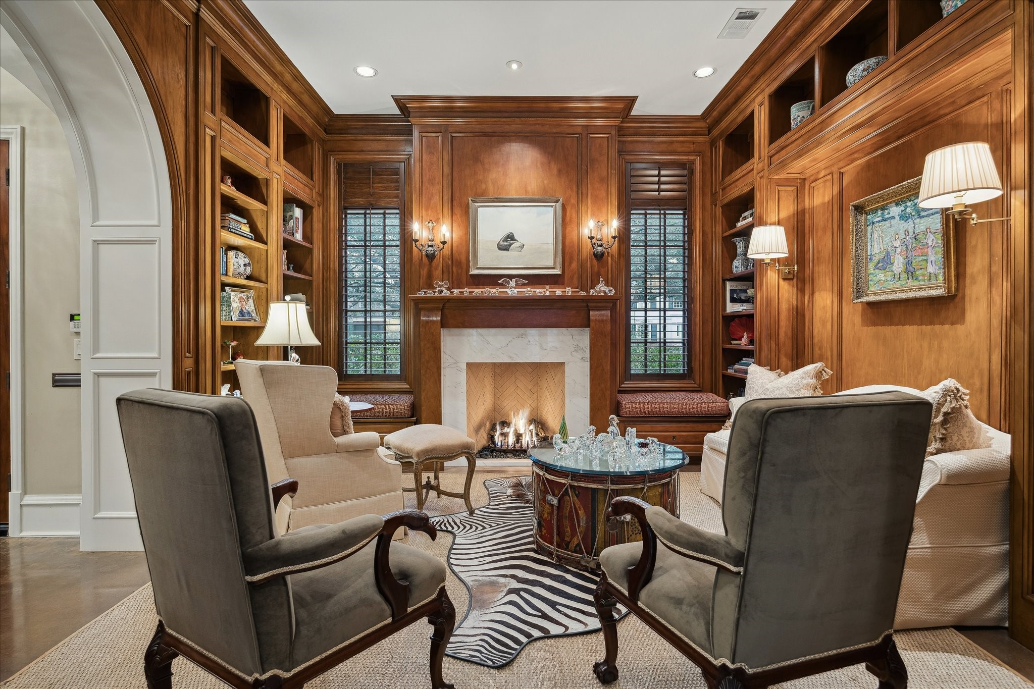3109 Locke Lane Houston, TX 77019 - Photo 5 of 50 A richly appointed library designed for both comfort and distinction, featuring custom built-in shelving and cabinetry framed by raised panel molding and warm wood paneling. A stone-surround fireplace with decorative iron screen anchors the room, while crown molding, arched entry, wall sconces, recessed lighting, and in-ceiling speakers create an inviting space bathed in natural light from thoughtfully placed windows.
