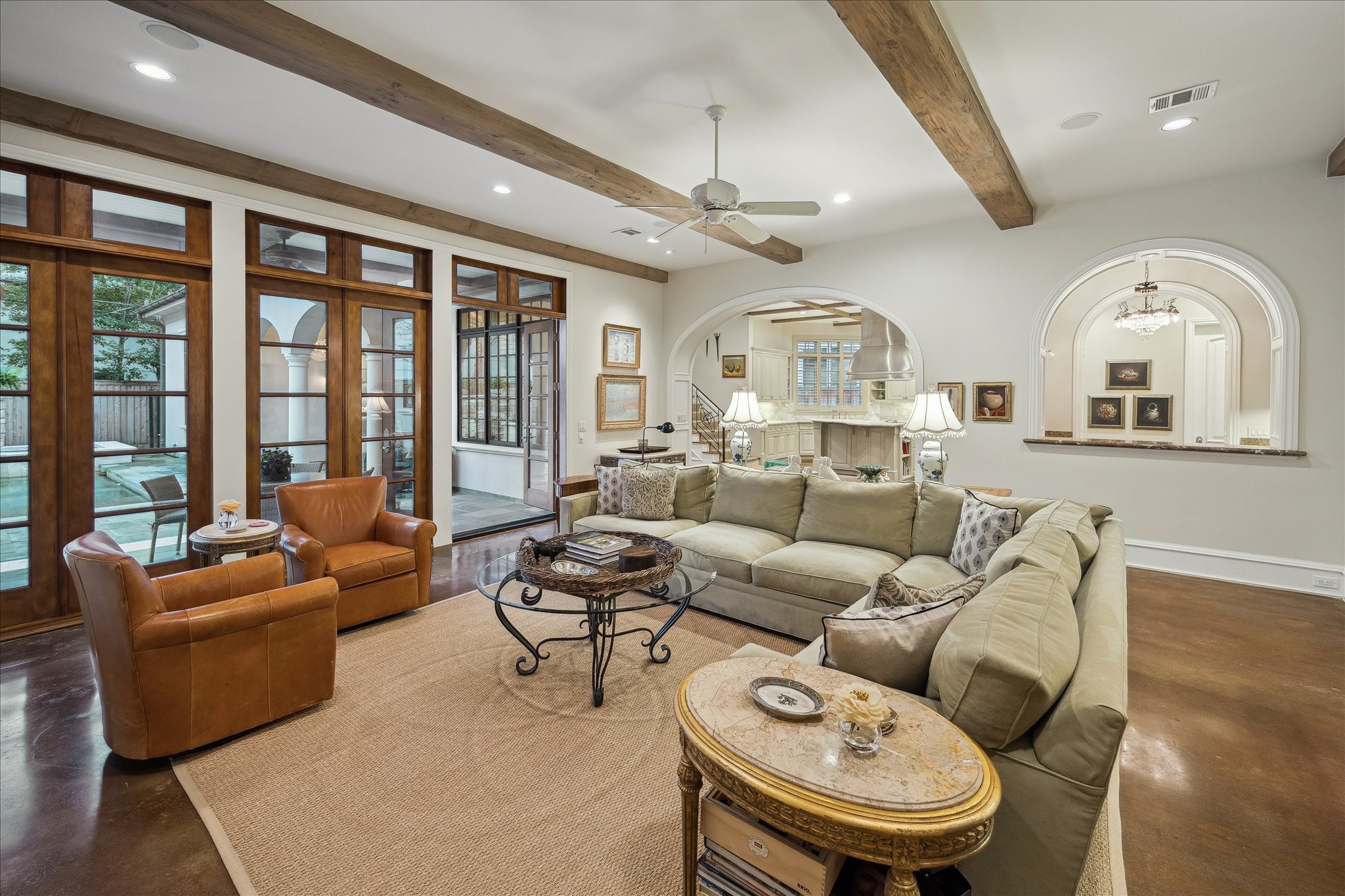 3109 Locke Lane Houston, TX 77019 - Photo 10 of 50 Exposed wood ceiling beams and polished concrete flooring set a refined yet relaxed tone in this expansive family room. A stone fireplace with decorative iron screen and herringbone detail serves as the focal point, complemented by custom shelving and abundant natural light from south-facing French doors overlooking the pool.