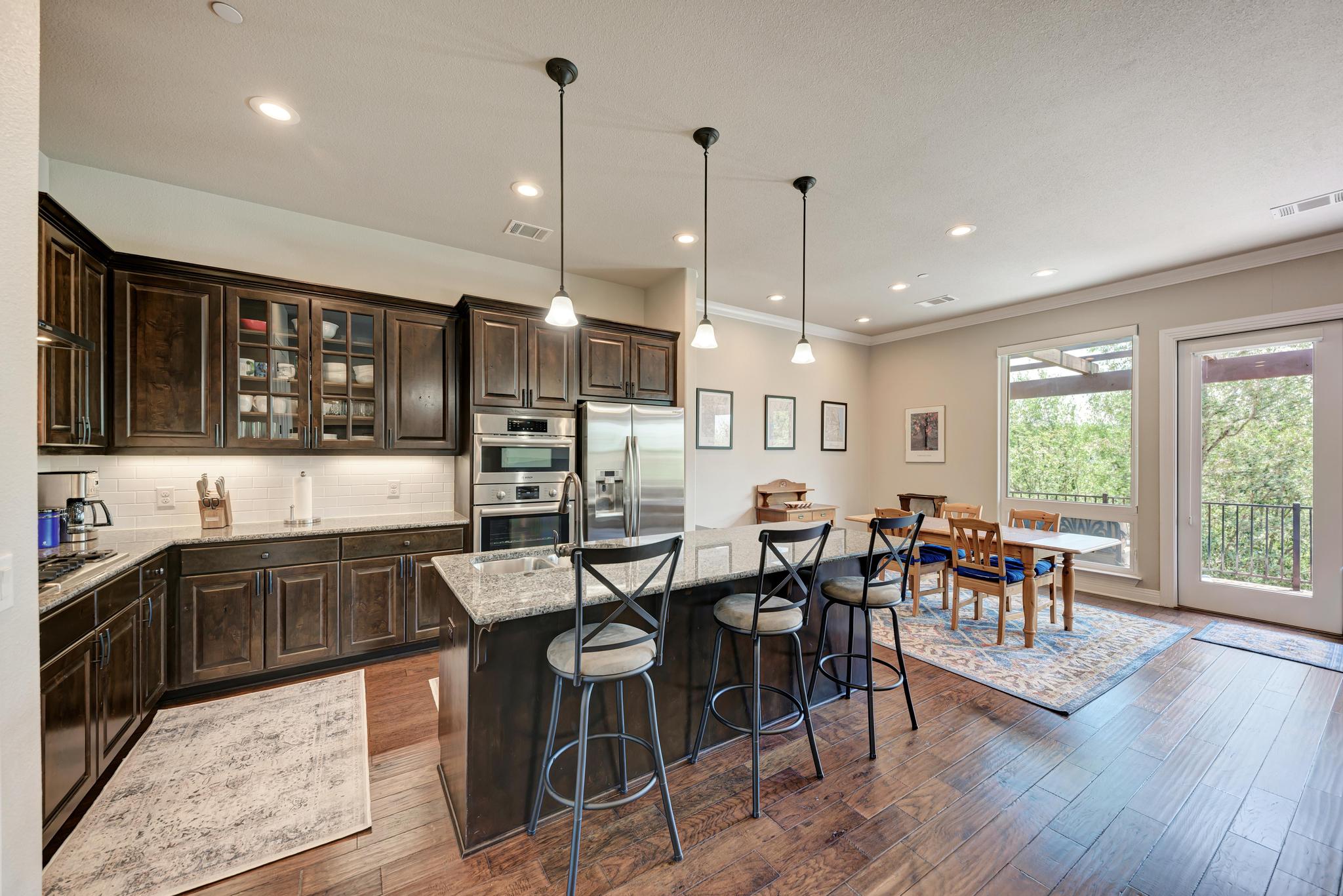 2601 North Quinlan Park Road, Unit 506 Austin, TX 78732 - Photo 12 of 39 An open concept kitchen features warm wood cabinetry, granite countertops, and stainless steel appliances, seamlessly flowing into a sunlit dining area.