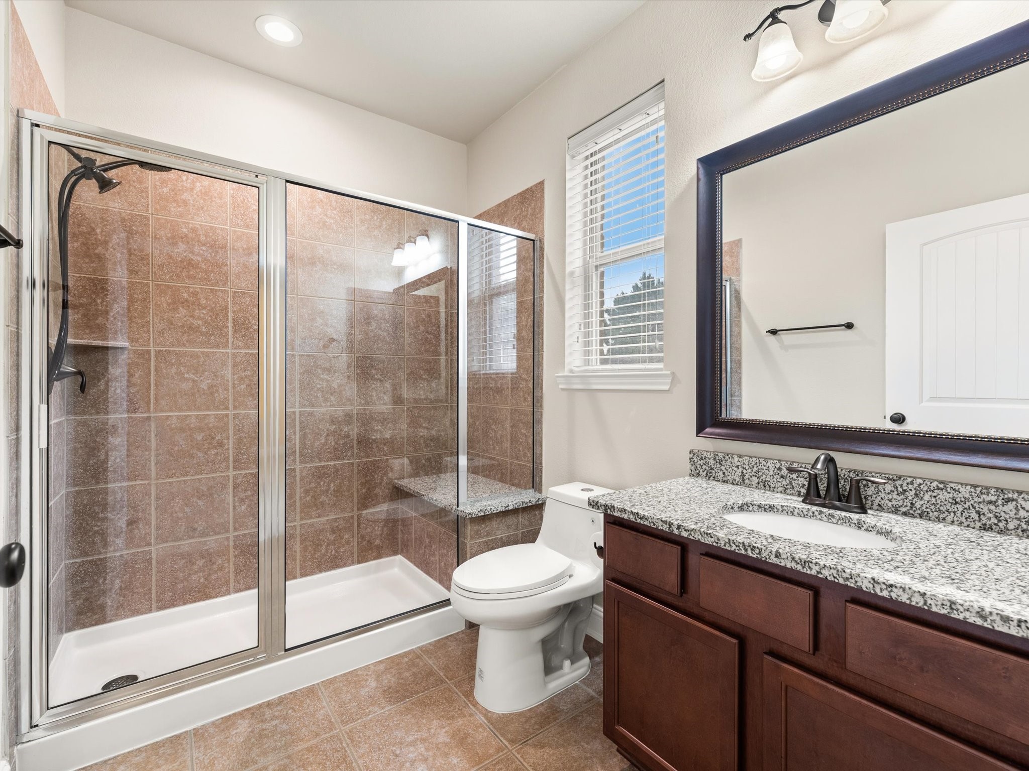 3907 Randle Ridge Court Fulshear, TX 77441 - Photo 25 of 46 This bathroom is inside the third first floor bedroom. It features a spacious glass-enclosed shower with a seat, a granite countertop with a single sink, dark wood cabinetry, and a large, framed mirror. There is a window for natural light, and the overall look is modern and clean.