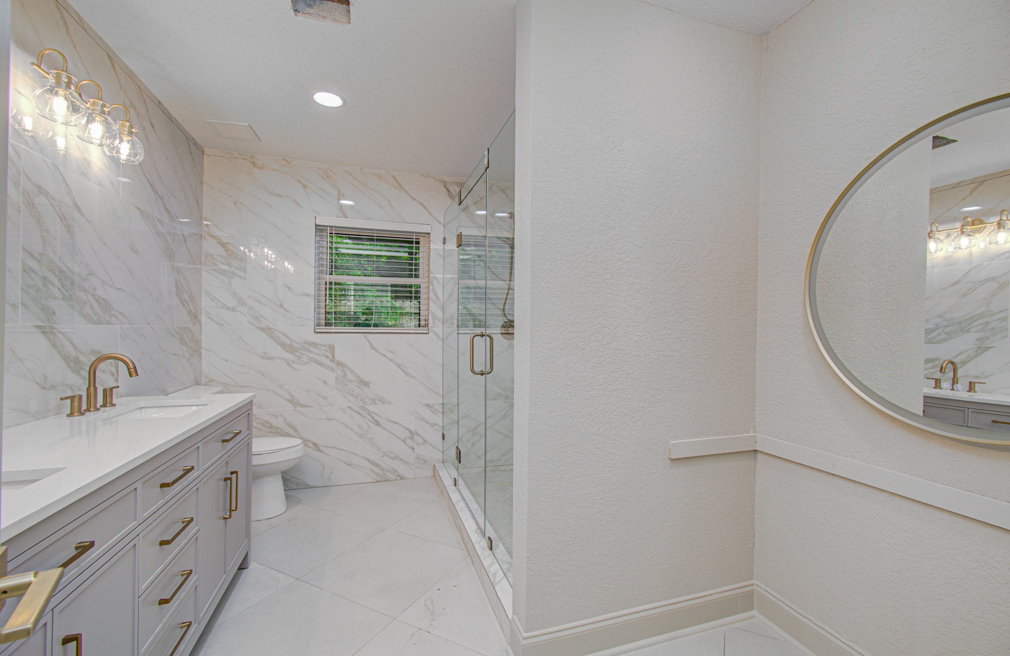 1801 Benbow Way Houston, TX 77080 - Photo 20 of 32 This modern bathroom features elegant marble walls, a spacious glass-enclosed shower, and a double vanity with gold fixtures. It includes a large round mirror and ample natural light from a window, creating a luxurious and inviting space.