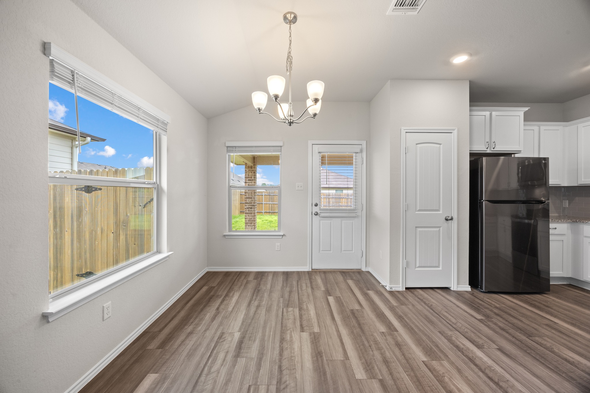 109 Gunther Way Taylor, TX 76574 - Photo 11 of 29 Open-concept dining area featuring wood-finish flooring, a contemporary chandelier, two windows with blinds, a back exterior door, and a partially visible kitchen with white cabinetry and a black refrigerator