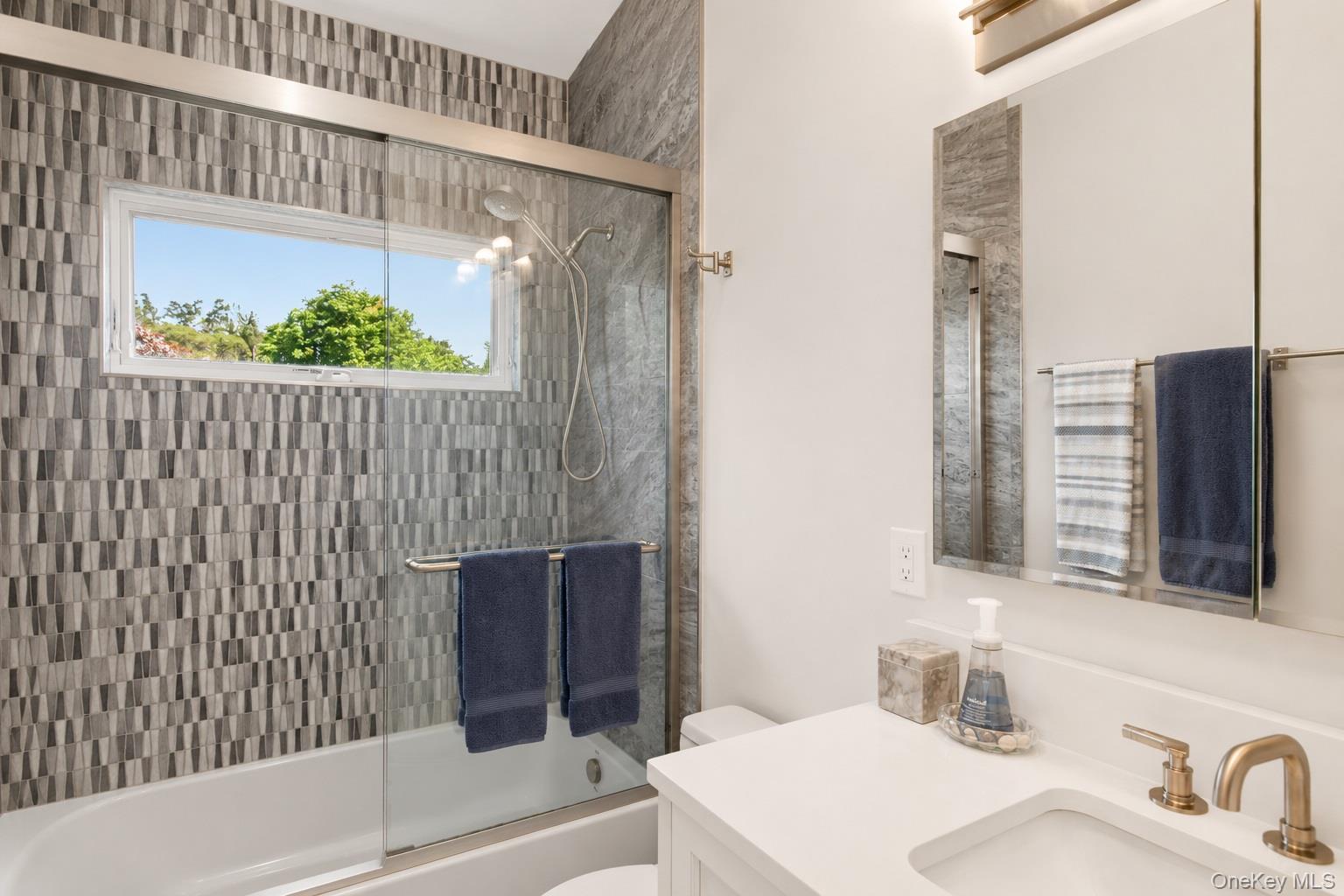 65 Shore Road Westhampton, NY 11977 - Photo 21 of 24 Contemporary bathroom featuring a combination shower and tub with sliding glass enclosure, mosaic-patterned tile surround, a horizontal window, and a single vanity with a white countertop and gold-toned fixtures