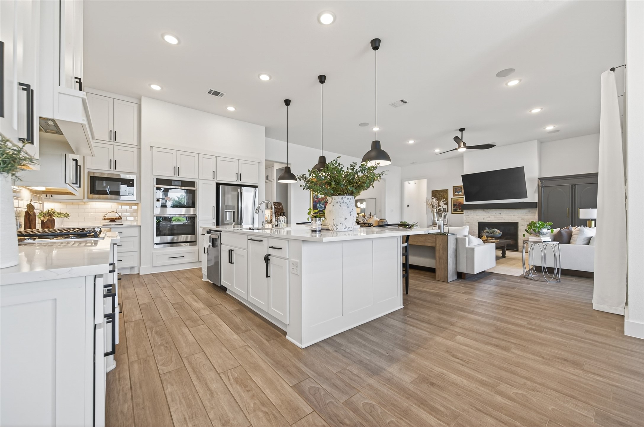 29612 Fulshear Lake Trace Fulshear, TX 77406 - Photo 26 of 48 This photo showcases a modern, open-concept kitchen and living area. The kitchen features a large island with pendant lighting, sleek white cabinetry, stainless steel appliances, and hardwood floors. The adjacent living space includes a cozy sitting area with a fireplace and a mounted TV, providing a comfortable and stylish environment.
