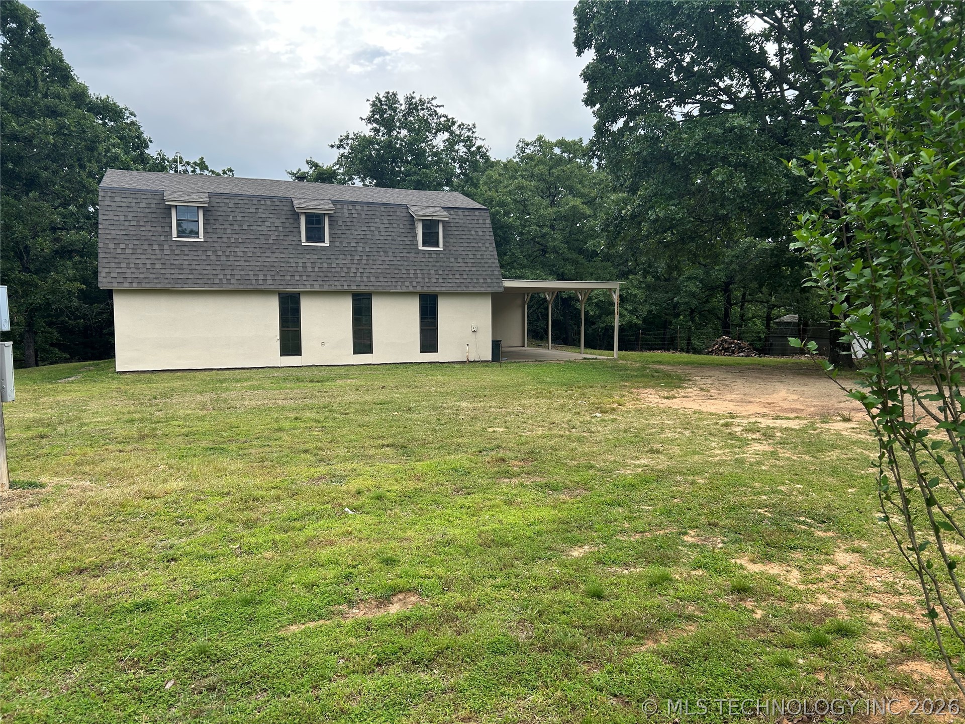 Gambrel roofline property featuring three dormer windows, a covered carport, and a surrounding natural landscape with mature trees