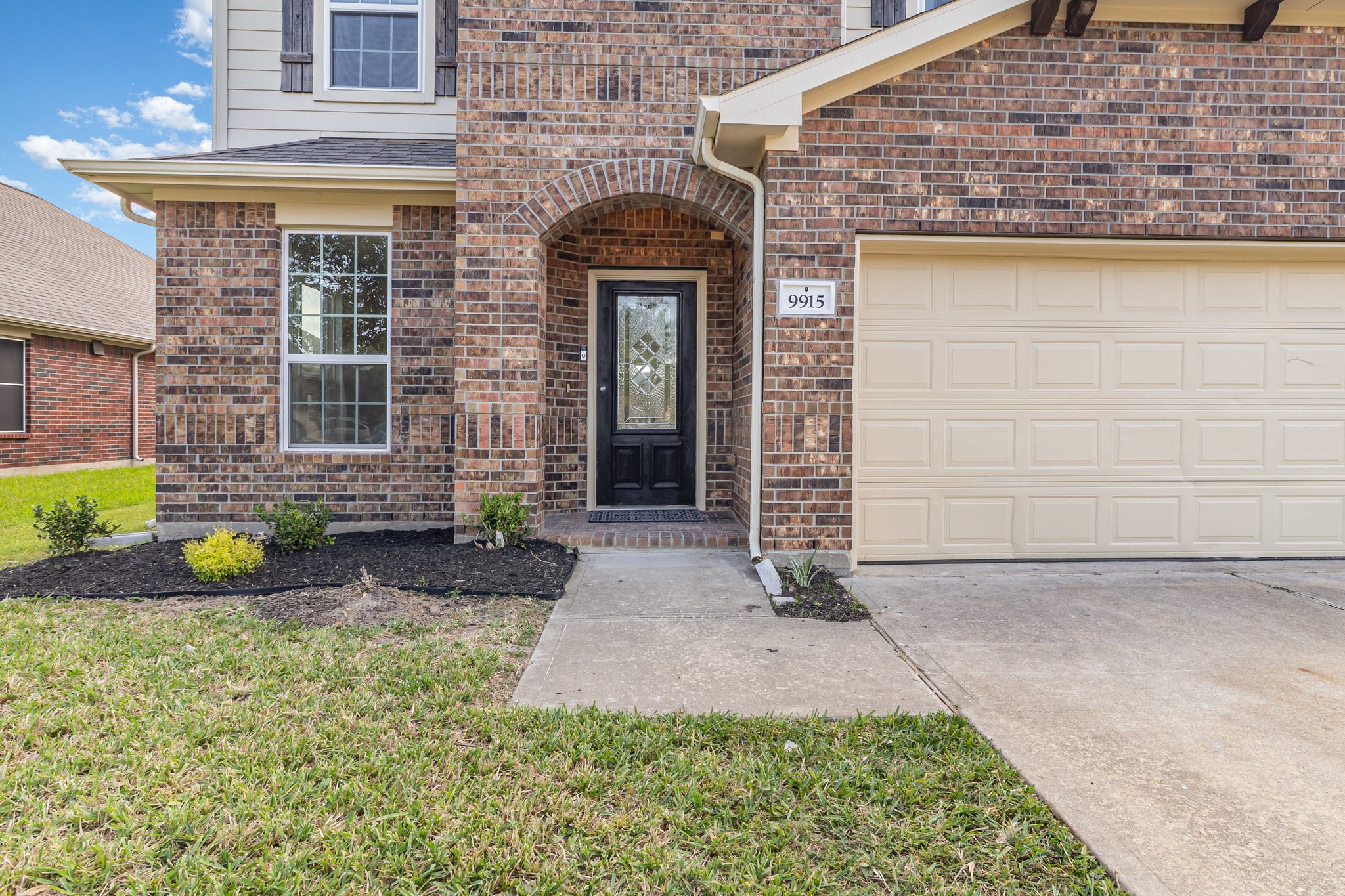 This photo shows a welcoming brick home exterior with a neatly landscaped front yard. It features a black front door under an arched entryway and an attached double garage.
