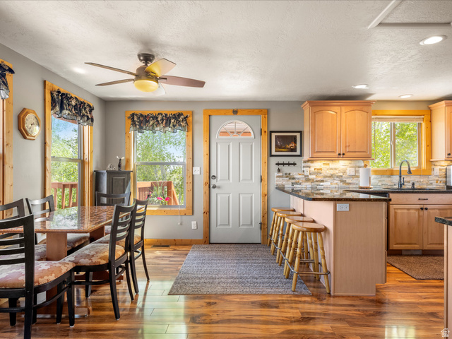 371 Lewis Loop, Unit 29 Fish Haven, ID 83287 - Photo 26 of 40 Open-concept kitchen and dining area featuring wood-finish flooring, light wood cabinetry, a granite countertop island with seating, a stone tile backsplash, and multiple windows with wood trim