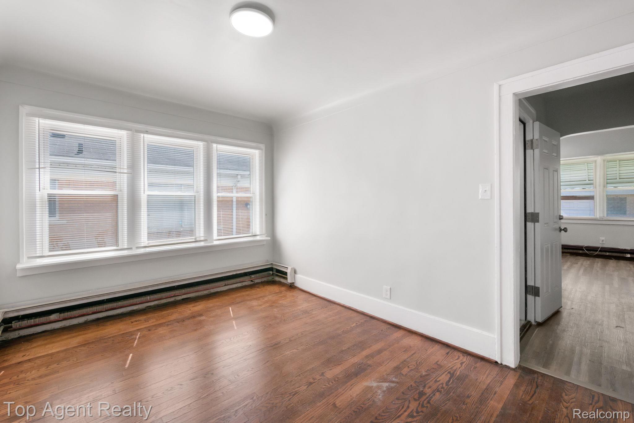 Wood-finish flooring with a white trim, featuring a three-panel window with horizontal blinds and a flush-mount ceiling light