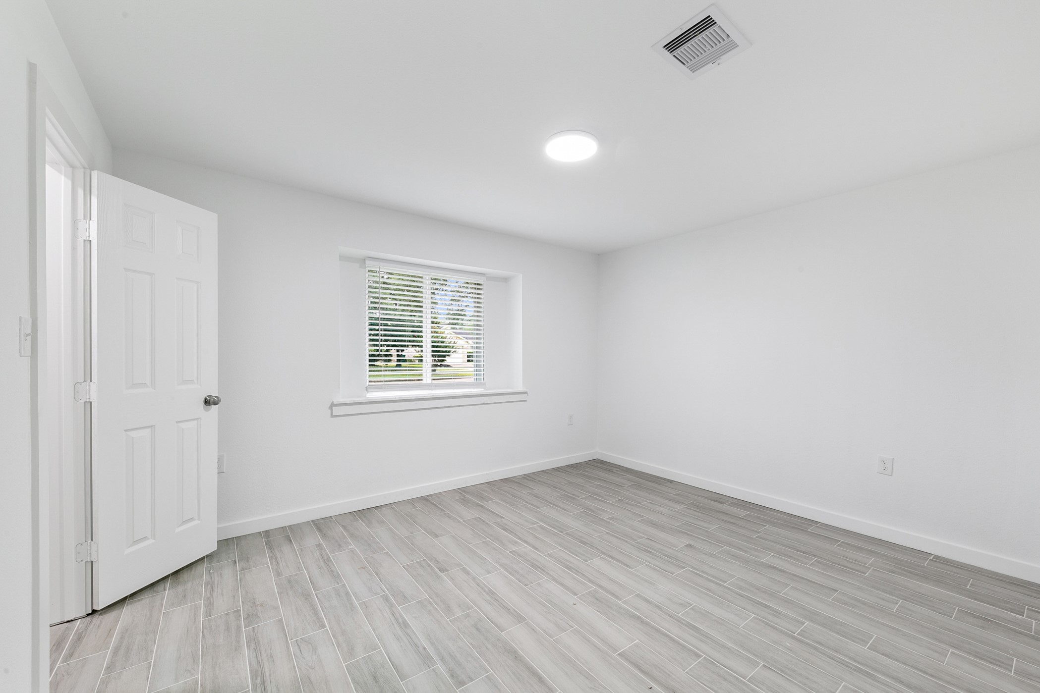 6811 McCullum Road Houston, TX 77489 - Photo 17 of 22 This photo showcases a bright, empty room with light gray wood-look tile flooring and white walls. It features a window with blinds, offering natural light, and an open door. The space is ideal for a bedroom or home office.