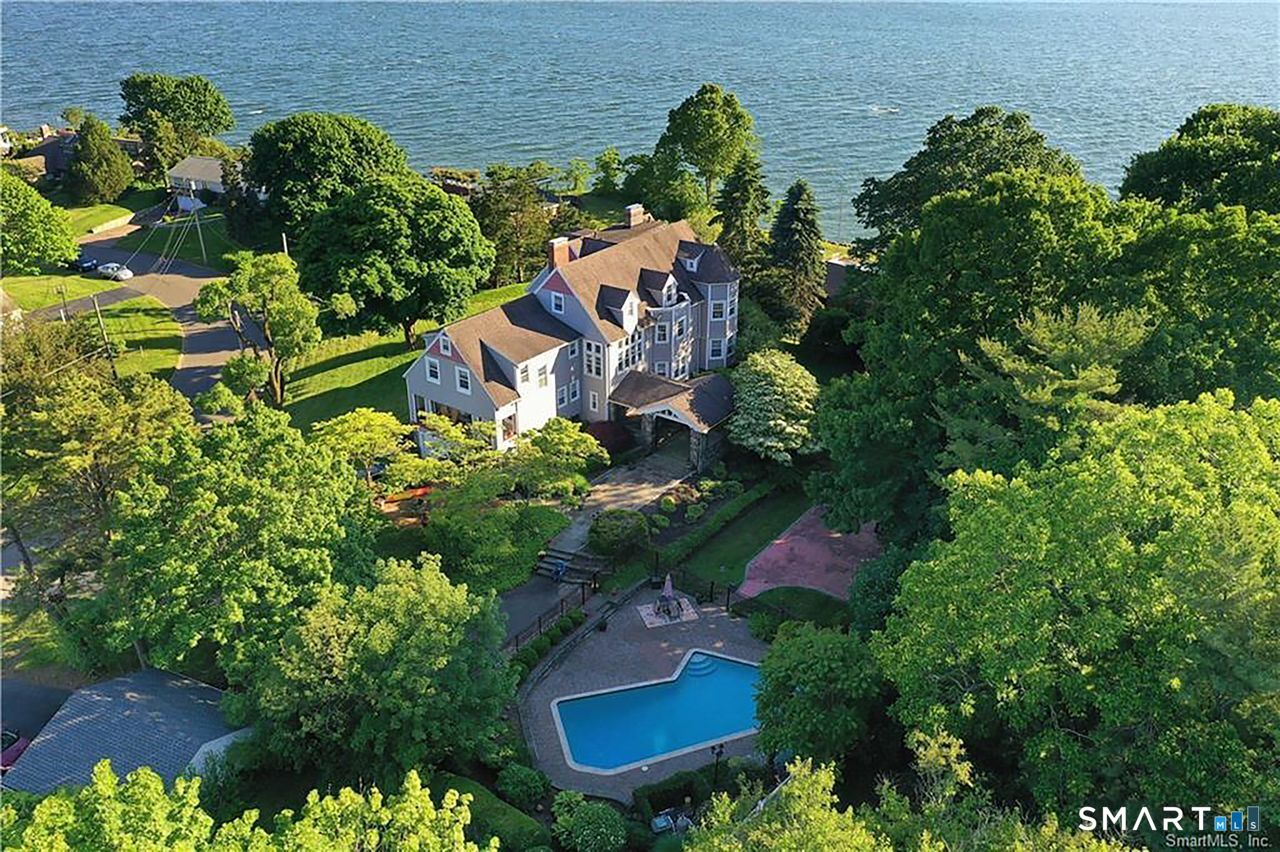 A breathtaking aerial view of the estate nestled among mature trees, showcasing its elevated setting, heated pool, and expansive grounds, all framed by sweeping vistas of Long Island Sound in three directions.
