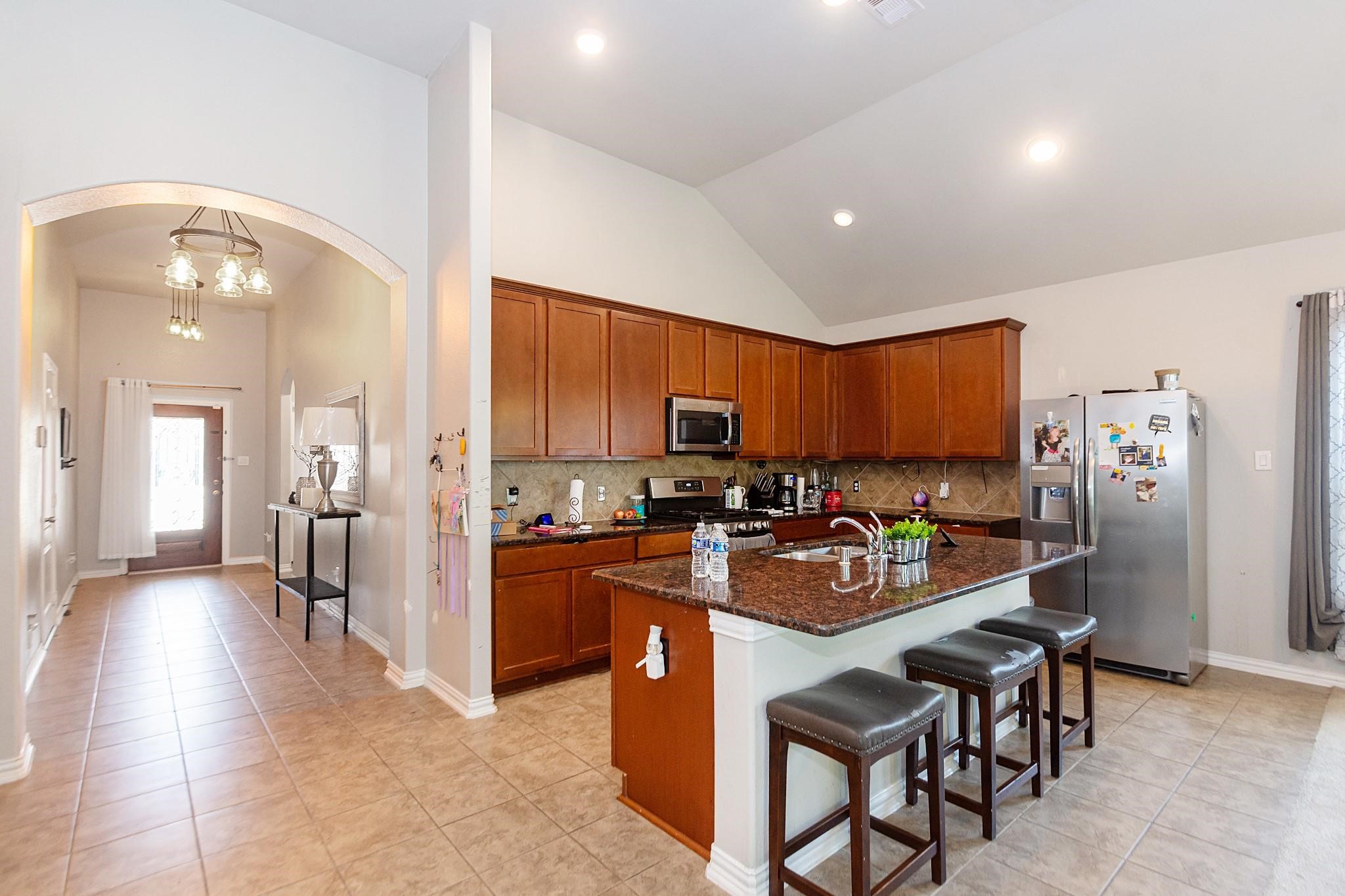 208 Timber Grove Court Clute, TX 77531 - Photo 5 of 22 This photo showcases a modern kitchen with warm wood cabinetry, granite countertops, and stainless steel appliances. The open layout includes a bar with seating and connects to a bright hallway with tile flooring, leading to a front door with glass panels for natural light.