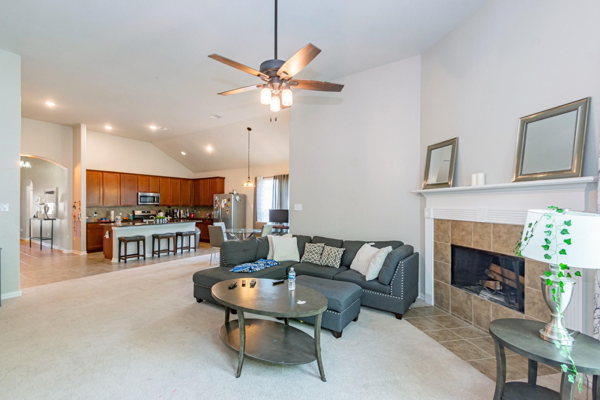 208 Timber Grove Court Clute, TX 77531 - Photo 9 of 22 This spacious living area features a high ceiling with a modern ceiling fan, a cozy fireplace, and an open-concept design that flows into the kitchen. The kitchen includes a large island with seating and stainless appliances, complemented by warm wooden cabinetry. The room is well-lit and inviting, ideal for entertaining or relaxing.