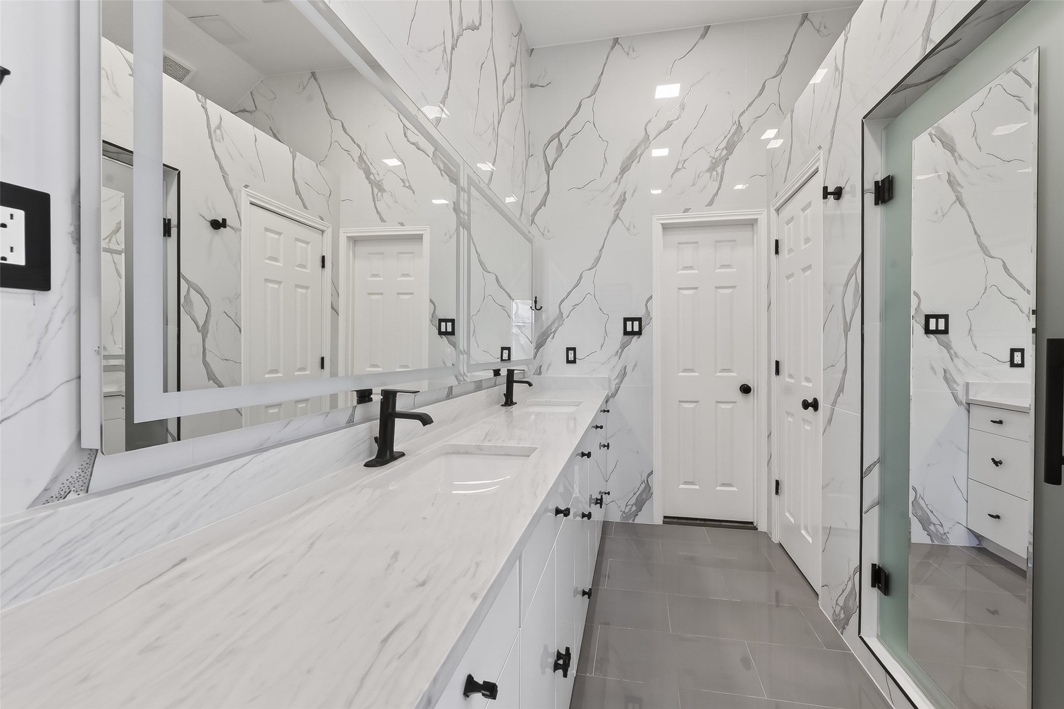 1311 Rene Court Sugar Land, TX 77479 - Photo 22 of 45 This primary bathroom features a sleek, modern design with marble-patterned walls, a long double vanity with black fixtures, and ample cabinetry. A large mirror enhances the space, complemented by elegant gray flooring.