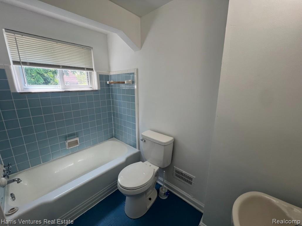 22148 Moross Road Grosse Pointe, MI 48236 - Photo 15 of 16 Bathroom featuring a white bathtub with blue tiled surround, a white toilet, and a window with horizontal blinds