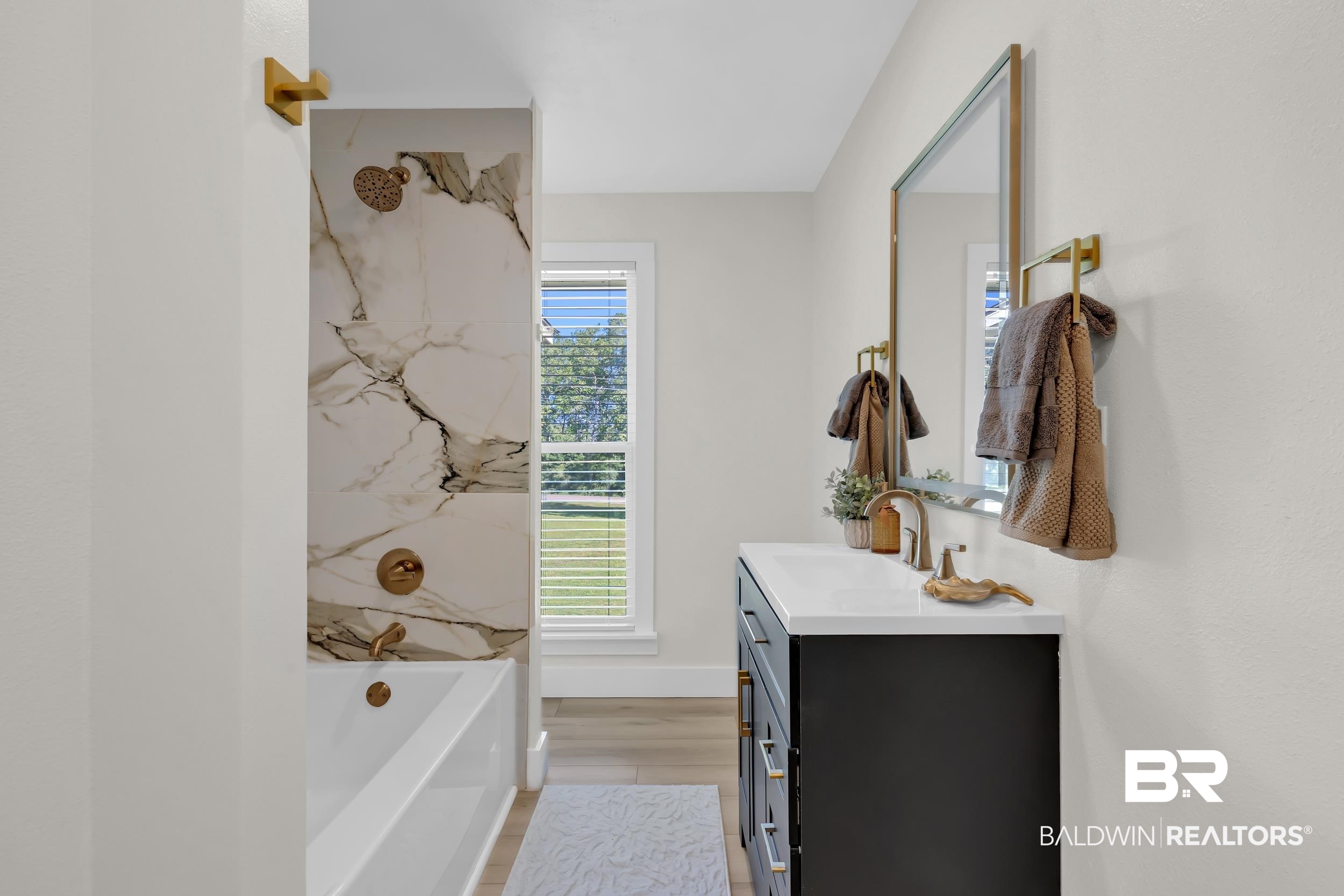 8170 Wilmer Georgetown Road Wilmer, AL 36587 - Photo 20 of 31 Modern bathroom featuring a freestanding tub with a partial marble-patterned tile wall, a black vanity with a white countertop, gold-finish fixtures, and light wood-finish flooring