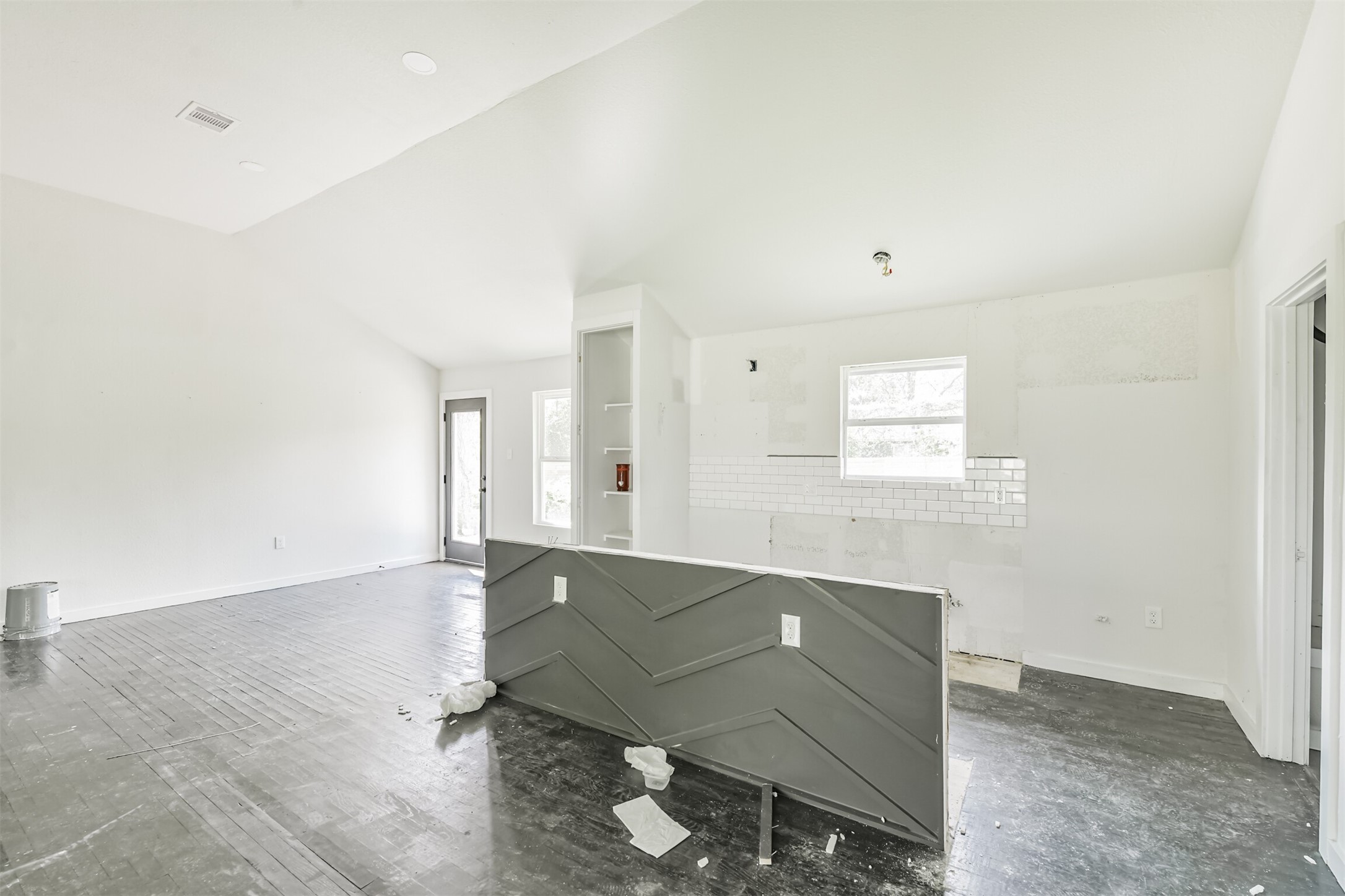 209 Leago Street Houston, TX 77022 - Photo 8 of 24 This photo shows a bright, open-concept space with high ceilings and potential for customization. The room features a partially finished kitchen area with white subway tiles and a gray partition wall, offering a modern touch. Large windows and a door provide ample natural light. Perfect for a buyer looking to personalize their home.