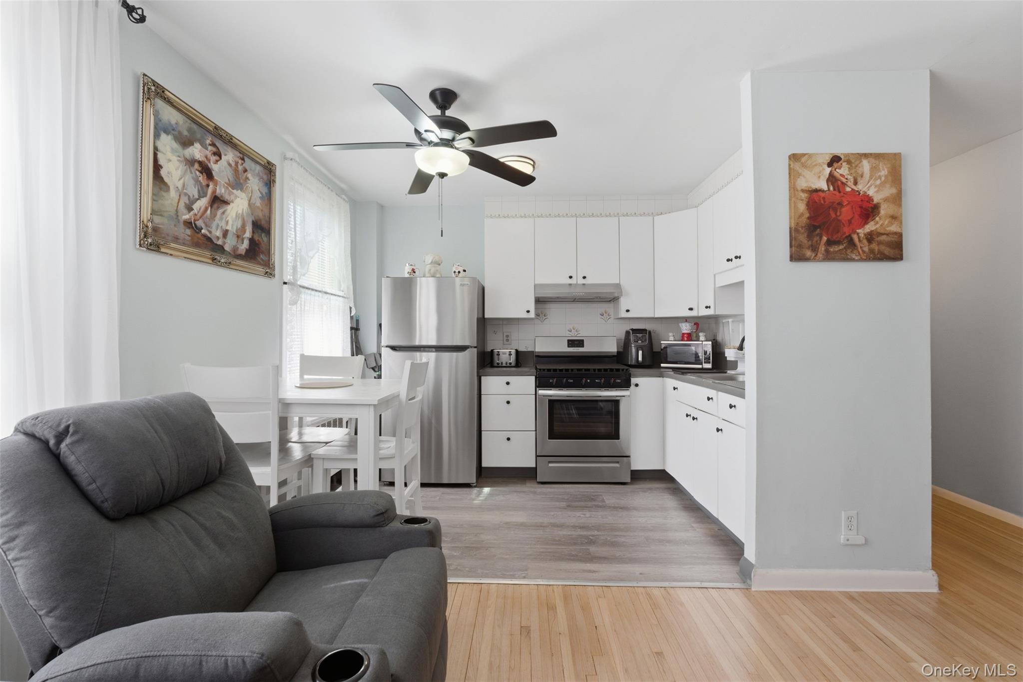 25 Trinity Place, Unit 2K New Rochelle, NY 10805 - Photo 9 of 24 The property features an open-concept living area with light hardwood flooring, a ceiling fan with integrated lighting, and a kitchen with white cabinetry and stainless steel appliances