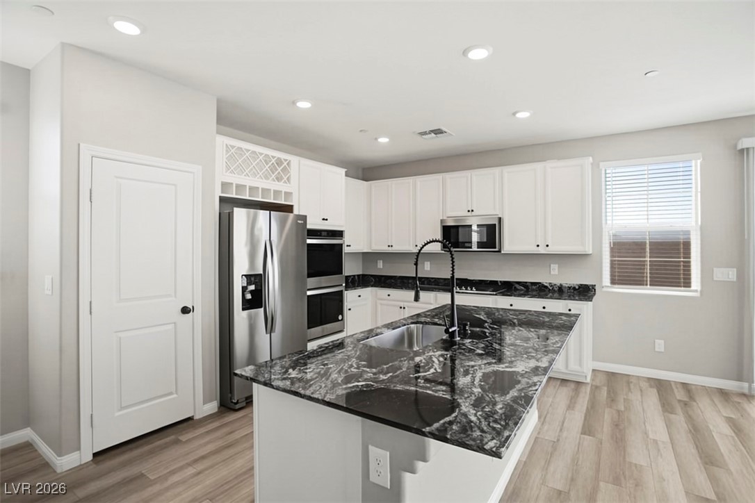 2865 Mincio Street Henderson, NV 89044 - Photo 7 of 33 Kitchen with dark stone countertops, appliances with stainless steel finishes, white cabinets, a center island with sink, and light wood-type flooring