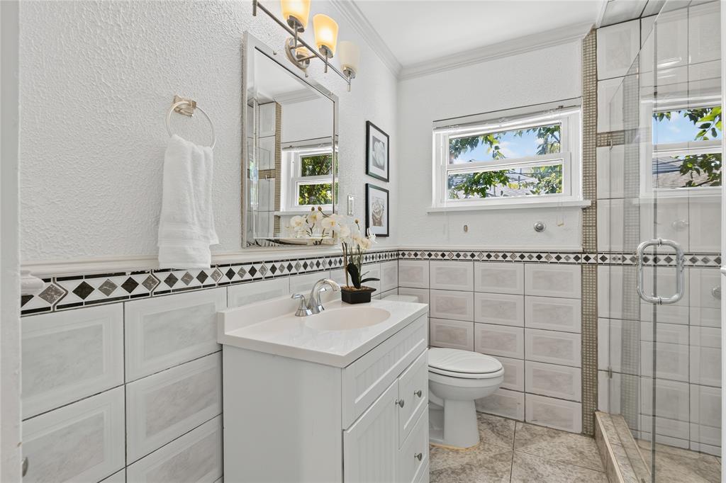 649 Peavy Road Dallas, TX 75218 - Photo 18 of 30 White vanity with countertop sink, framed mirror with sconce lighting, tiled walls with decorative border, and a glass-enclosed shower