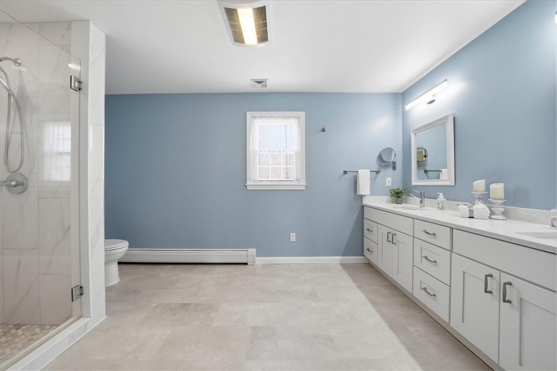 32 Lisa Lane Hopkinton, RI 02832 - Photo 31 of 50 This is not your typical ensuite—this is a massive, fully renovated primary bath that truly stands out. Featuring an extended double vanity w/ quartz countertops, designer lighting & incredible storage, plus a sleek tiled walk-in shower w/ glass enclosure. Added luxuries like a heated towel rack & heat lamp elevate the space even further. With its impressive scale & spa-like feel, this bathroom is a rare & unforgettable highlight of the home.