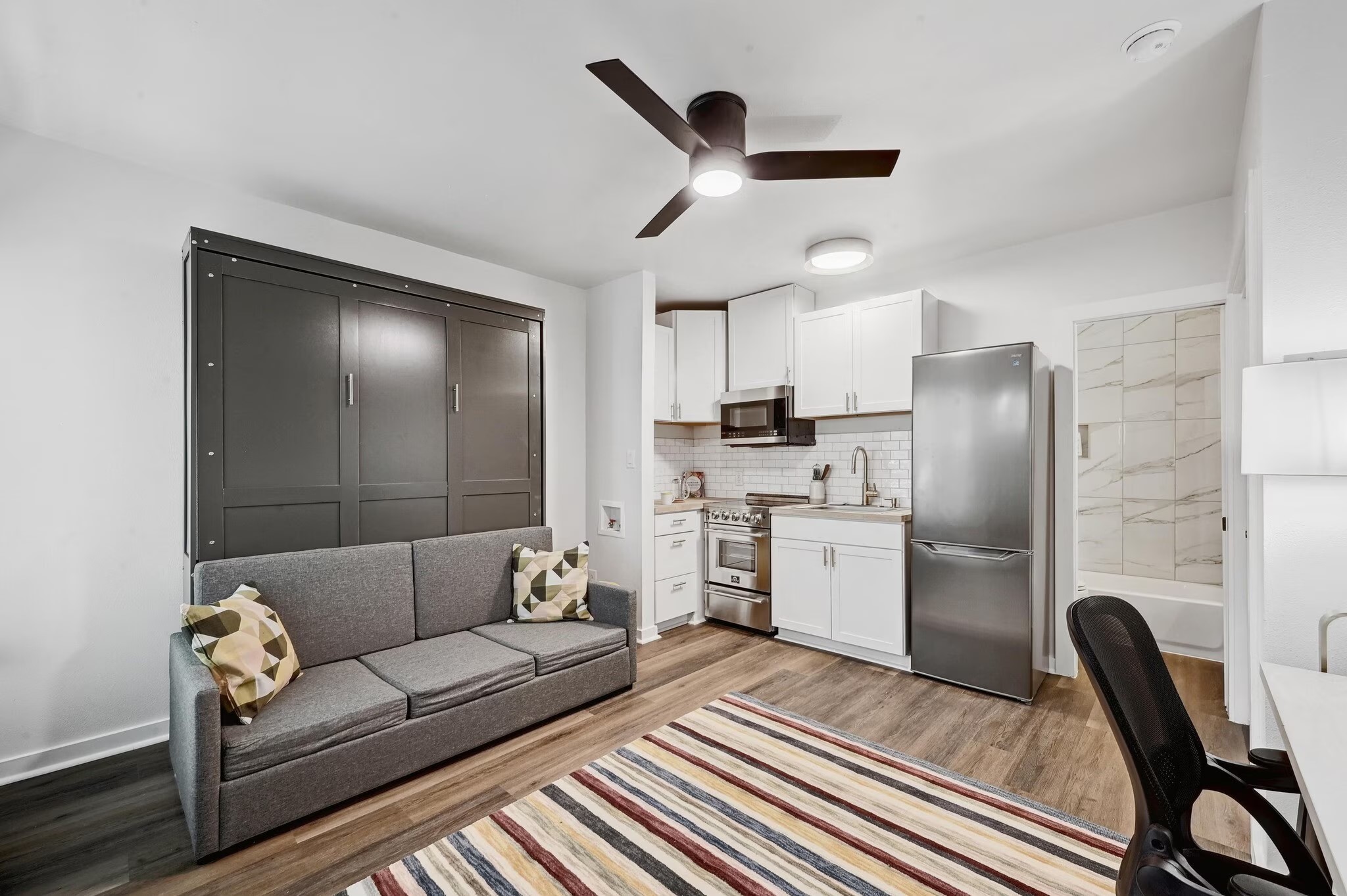 2802 Whitis Avenue, Unit 106 Austin, TX 78705 - Photo 1 of 8 Open-concept living area featuring wood-finish flooring, a contemporary ceiling fan with integrated lighting, and a kitchen with white cabinetry, stainless steel appliances, and a subway tile backsplash