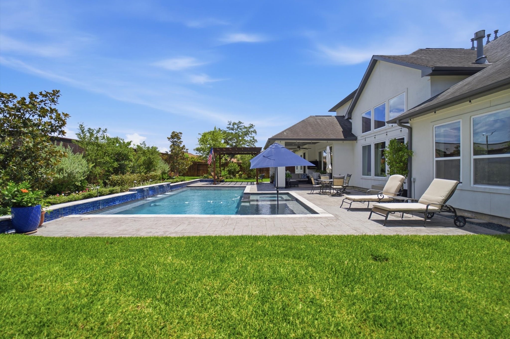 23722 Thortons Park Lane Spring, TX 77389 - Photo 46 of 48 This photo showcases a modern backyard with a spacious in-ground pool, surrounded by a well-maintained patio and greenery. The house features large windows and a covered seating area, ideal for outdoor relaxation and entertaining.