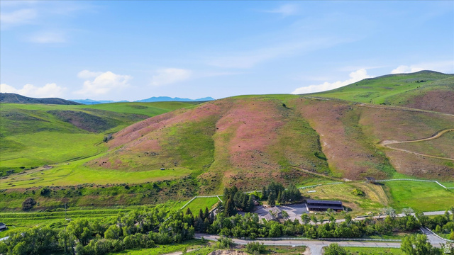 2075 Canyon Road Smithfield, UT 84335 - Photo 4 of 18 Expansive rural landscape featuring rolling green and brown hills, a dark-roofed building surrounded by trees, and a paved road with white fencing