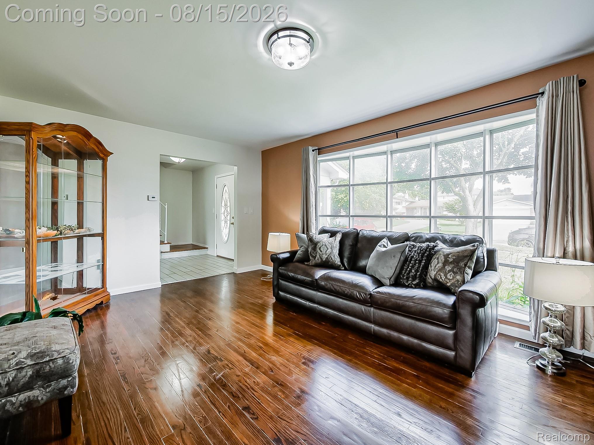 21131 Harvard Road Southfield, MI 48076 - Photo 3 of 33 Spacious living room showcasing rich wood-finish flooring, a black leather sofa with decorative pillows, and an expansive multi-pane window wall with thermal-lined drapery