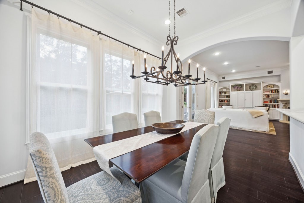 2508 Brun Street Houston, TX 77019 - Photo 12 of 50 This photo showcases a bright, open-concept dining and living area. It features large windows with sheer curtains, a stylish chandelier over a dining table, and an inviting living space with built-in shelving and ample lighting. The dark wood floors add a touch of elegance.