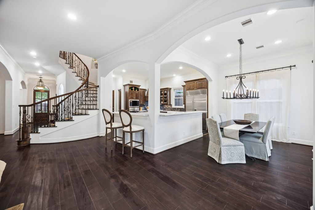2508 Brun Street Houston, TX 77019 - Photo 10 of 50 This photo showcases a spacious, elegant interior. It features an open layout connecting a dining area and kitchen, with a stylish chandelier and modern appliances. A grand, curved staircase with ornate railing adds sophistication, leading to an upper level, enhancing the home's luxurious feel.