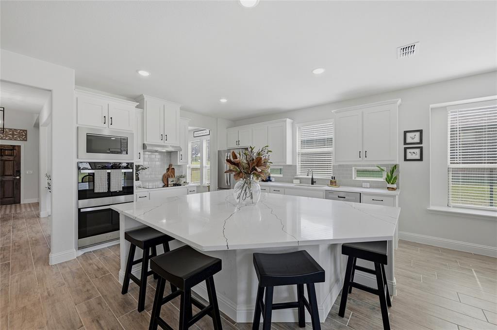 864 Shadow Valley Road Whitesboro, TX 76273 - Photo 11 of 40 Spacious kitchen featuring an oversized island with countertop seating, white shaker cabinetry, stainless steel built-in appliances, a subway tile backsplash, and wood-finish flooring