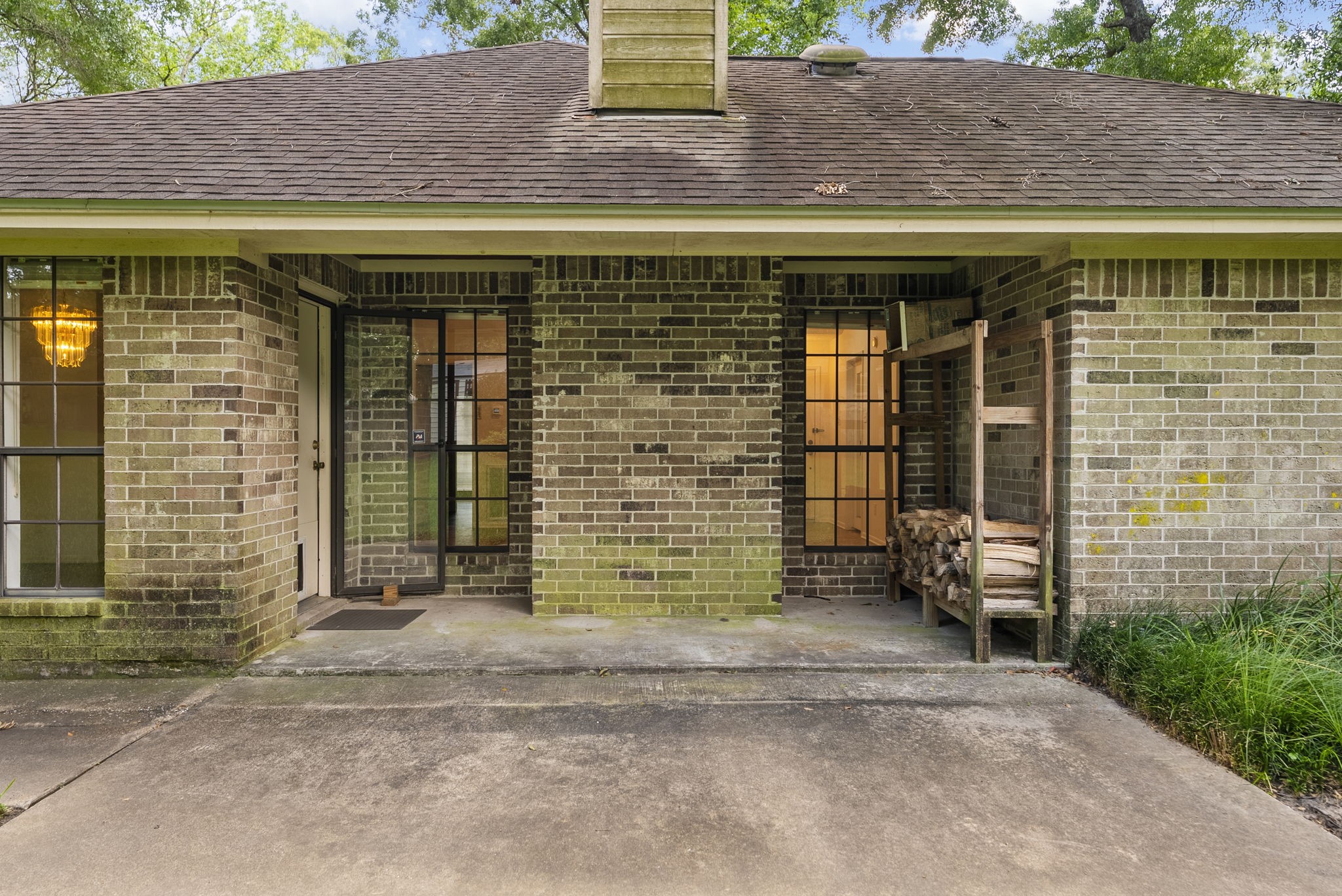 202 Sunset Boulevard Conroe, TX 77303 - Photo 41 of 50 This photo showcases a charming brick entryway with a covered porch, featuring large windows and a space for firewood storage. The area is surrounded by greenery, providing a welcoming and cozy atmosphere.