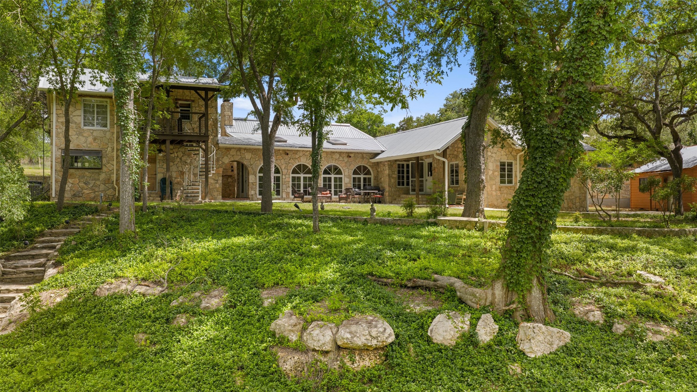 505 Red Corral Ranch Road Wimberley, TX 78676 - Photo 21 of 40 A stone-clad home nestled among mature trees, featuring a metal roof and a spiral staircase to an upper balcony.