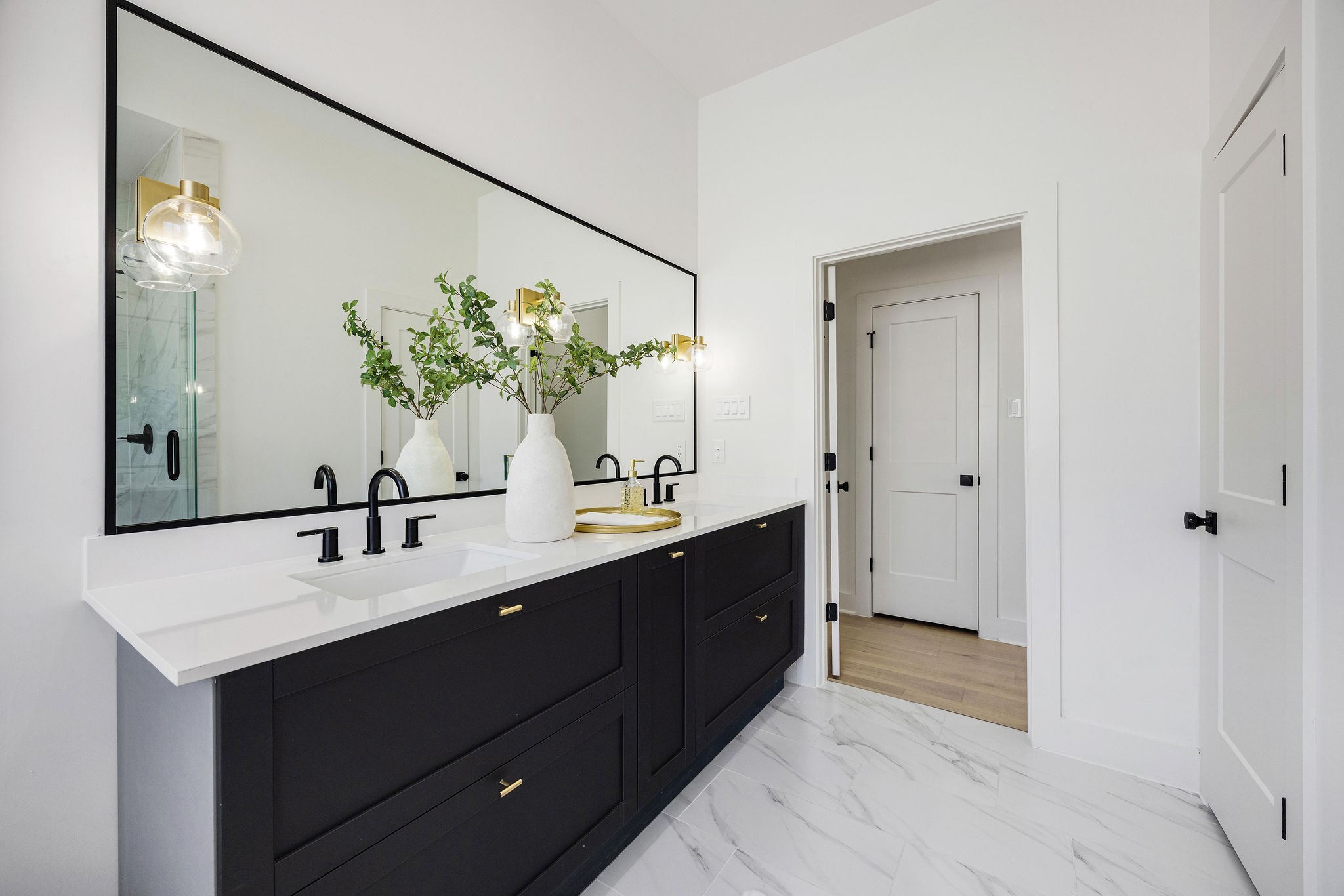 303 Marcella Street, Unit P Houston, TX 77091 - Photo 15 of 26 This modern bathroom features a sleek double vanity with black cabinets and brass accents, a large framed mirror, and stylish lighting. The white walls and marble-like flooring create a clean, sophisticated look.