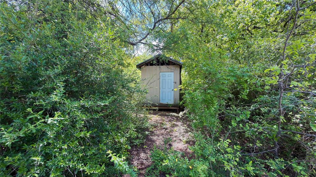 2900 Robinson Road Denton, TX 76210 - Photo 26 of 33 Freestanding utility building with a light blue door and wood siding, surrounded by mature trees and abundant shrubbery
