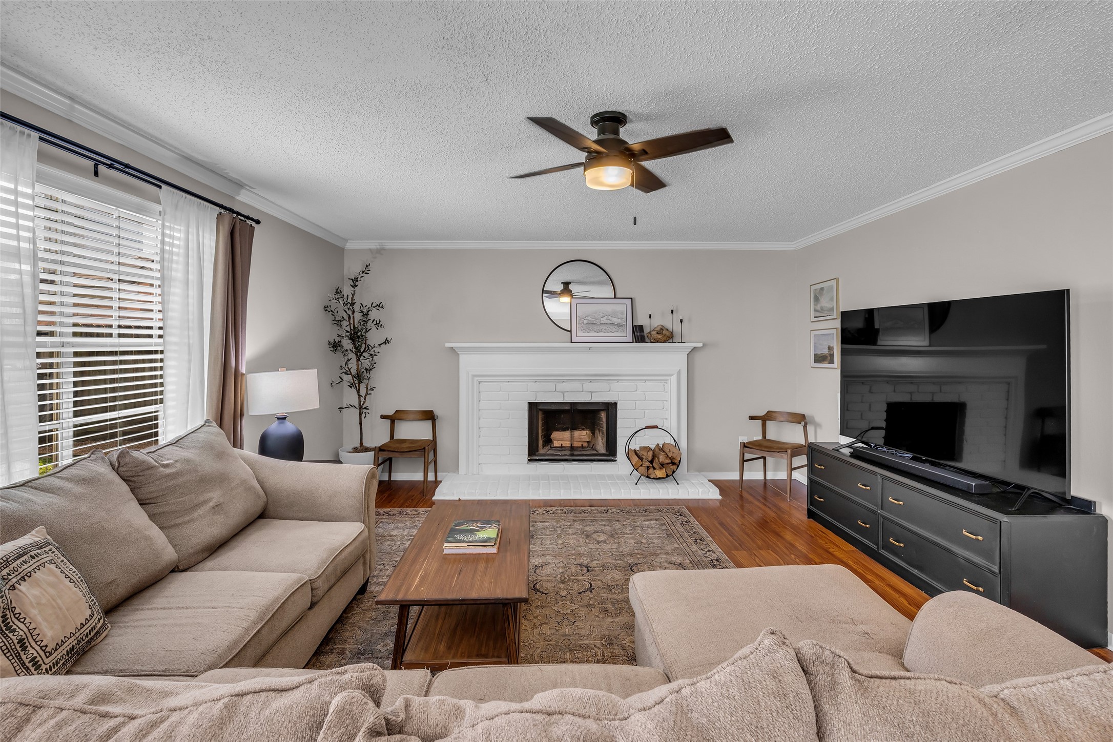 9415 Towerstone Drive Spring, TX 77379 - Photo 2 of 44 This cozy living room features a comfortable sectional sofa, a modern ceiling fan, and a large TV. The room is anchored by a white brick fireplace with a simple mantel, adorned with decorative items. Natural light streams through the large windows with elegant curtains, highlighting the warm hardwood floors.