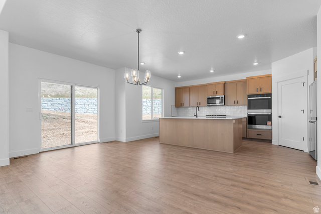 1341 West 2350 South, Unit 10 Perry, UT 84302 - Photo 5 of 18 Open-concept living space featuring wood-finish flooring, a modern kitchen island, light wood cabinetry, white subway tile backsplash, and stainless steel appliances including a double oven and microwave