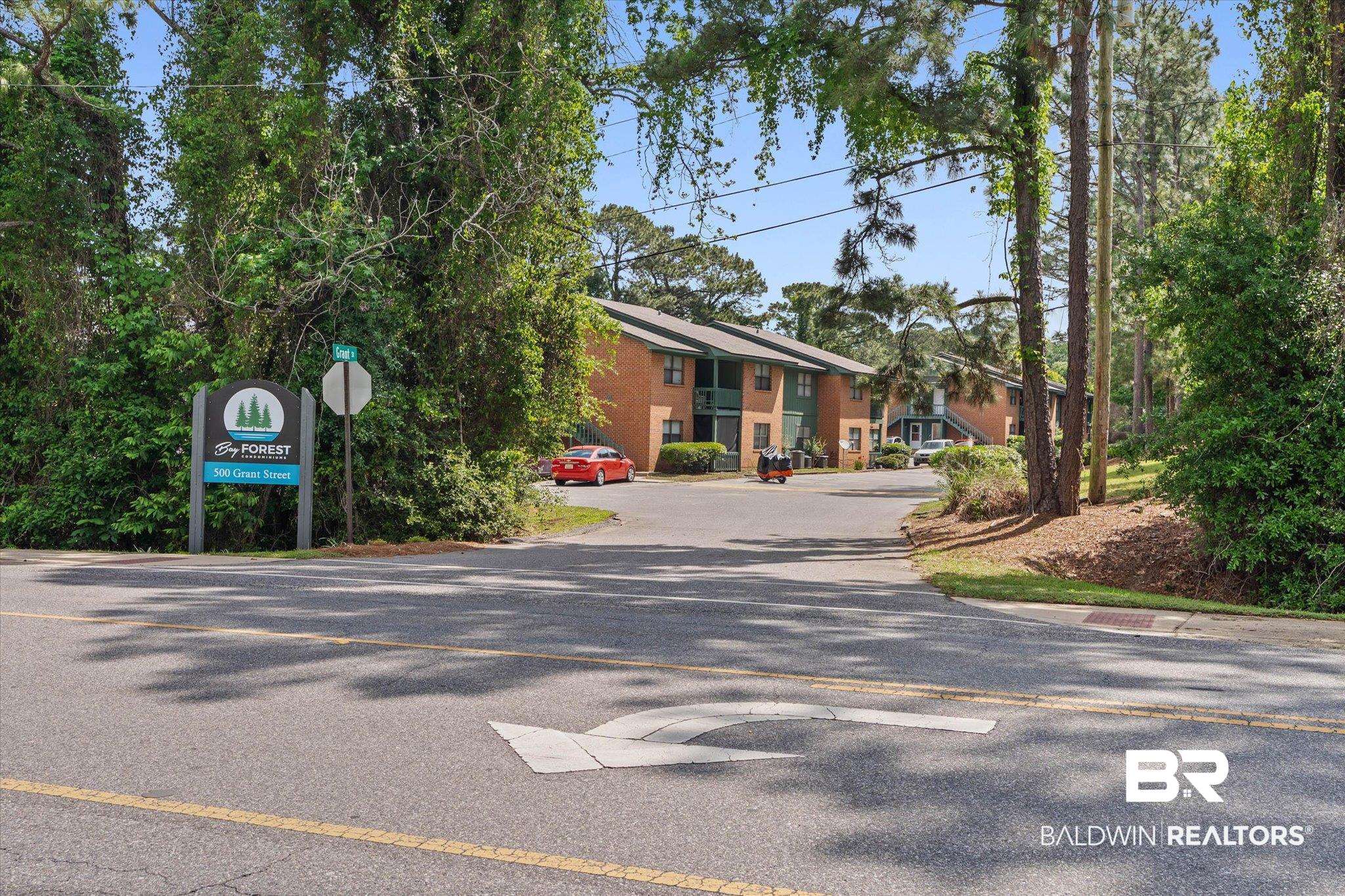 500 Grant Street, Unit D213 Daphne, AL 36526 - Photo 3 of 6 Paved road entrance to a property featuring an apartment complex sign framed by mature trees and shrubs