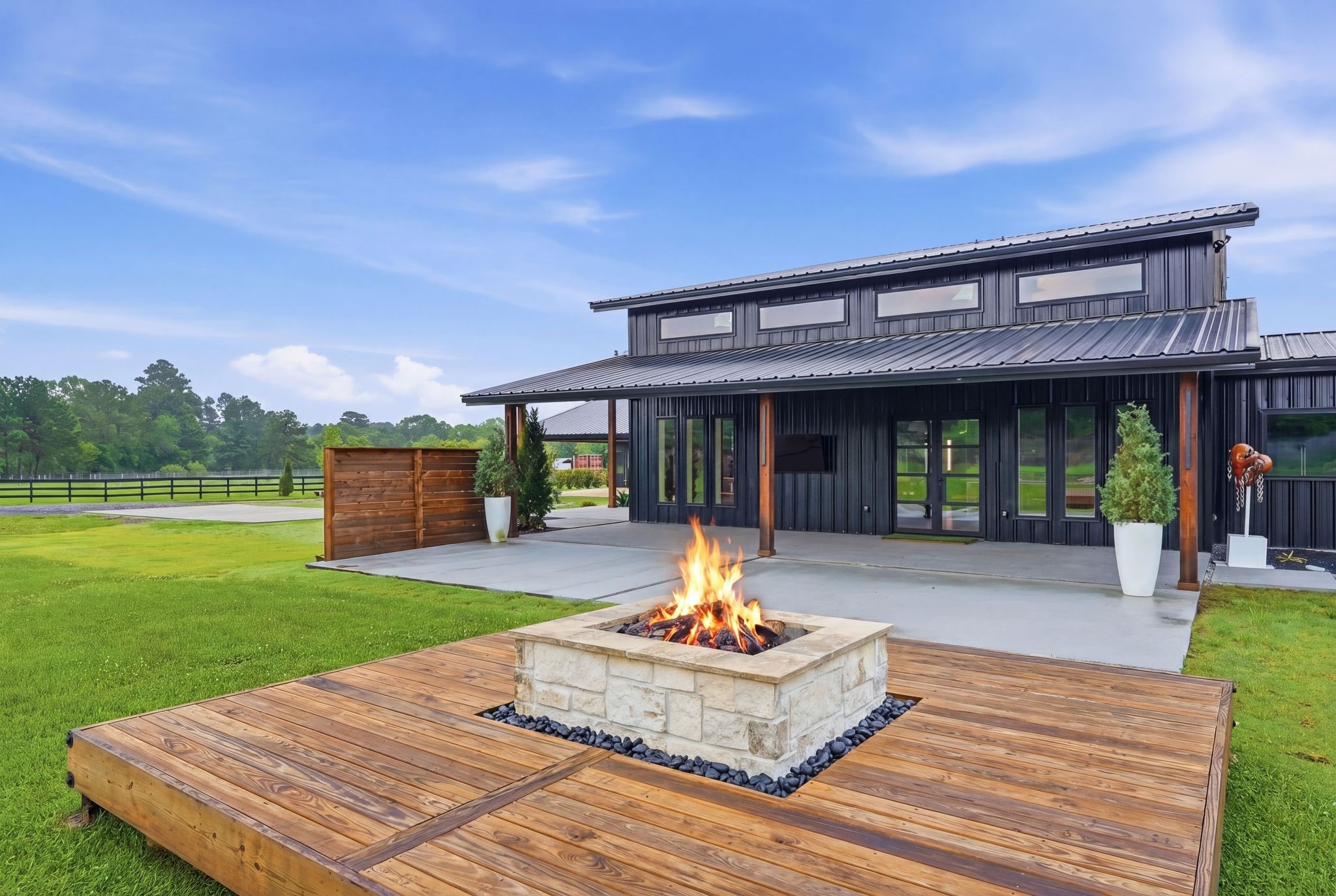 2743 Bobville Road Montgomery, TX 77316 - Photo 46 of 50 Modern home with a sleek black exterior and large windows. The backyard features a spacious patio with a built-in fire pit on a wooden deck, surrounded by lush green lawn and scenic views.