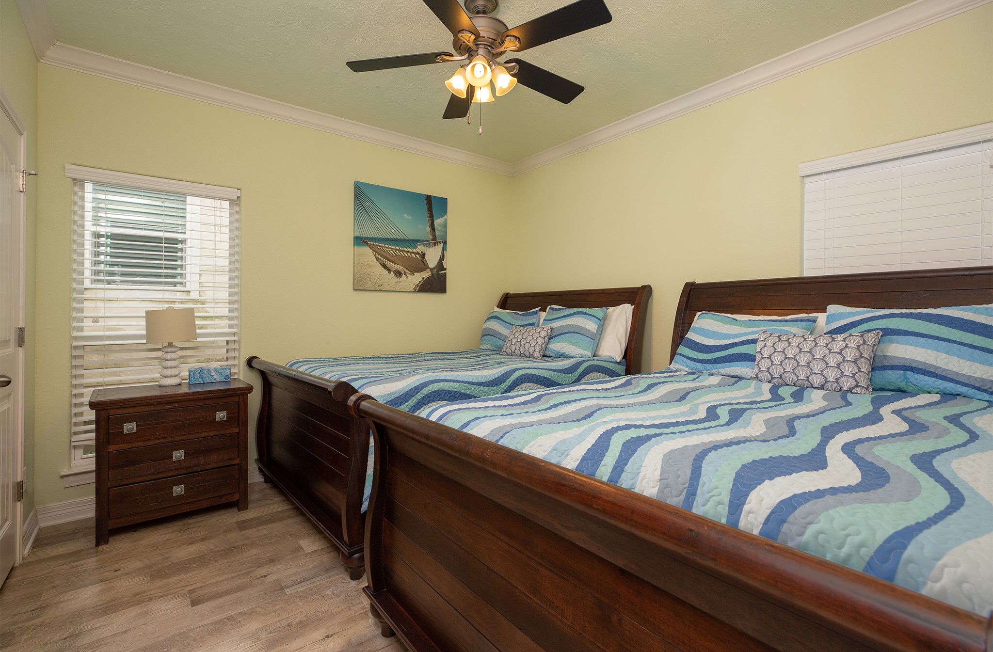 843 South Jacks Road Crystal Beach, TX 77650 - Photo 40 of 45 This cozy bedroom features two queen sized beds with vibrant blue and green bedding, a ceiling fan, and a window with blinds, offering a bright and inviting atmosphere. A small dresser with a lamp adds functionality, and the walls are a soft, calming color.
