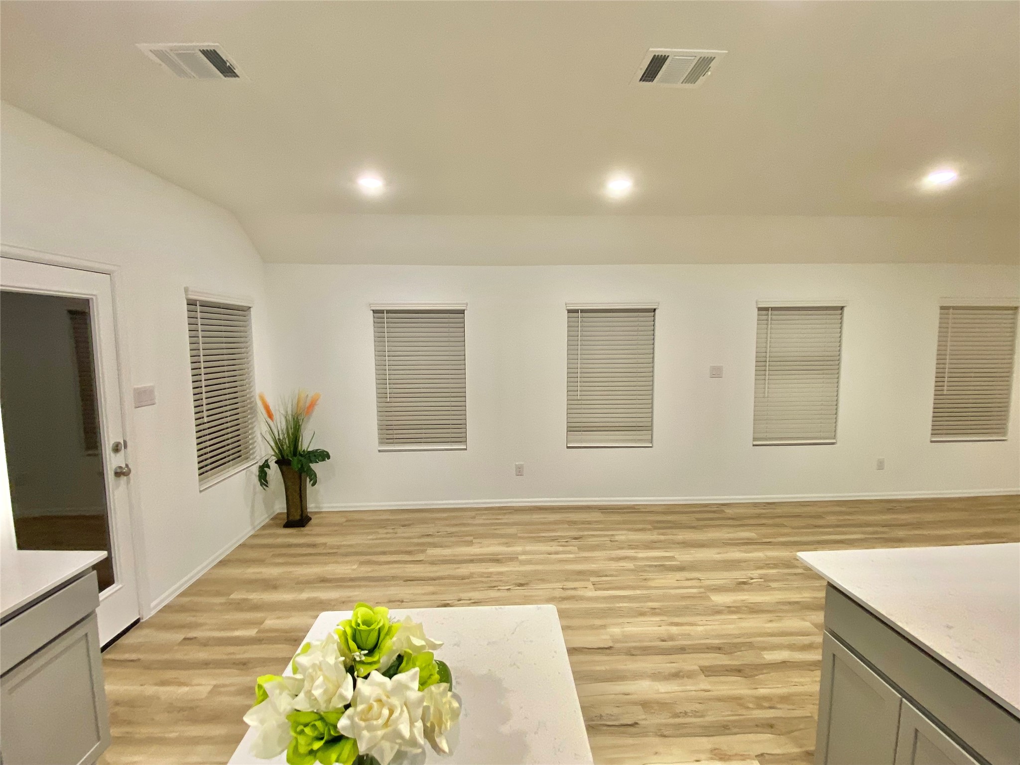 3724 Moss Hl Road Rosenberg, TX 77469 - Photo 13 of 29 This spacious, open-plan room features light wood flooring, recessed lighting, and ample windows with blinds. A modern kitchen island with a floral centerpiece adds elegance, and a door leads outside. Perfect for entertaining and relaxation.