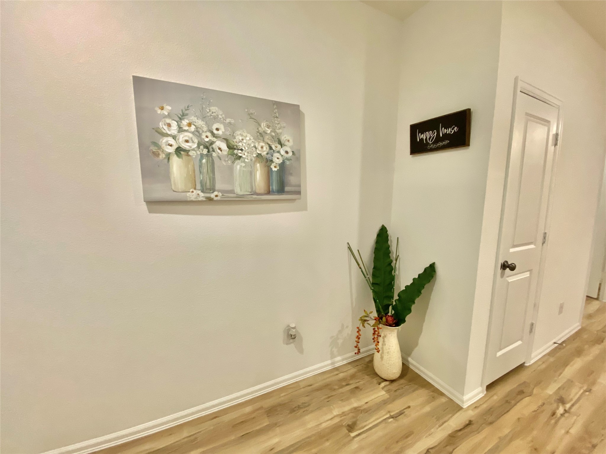 3724 Moss Hl Road Rosenberg, TX 77469 - Photo 3 of 29 This photo shows a bright hallway with light wood flooring. The walls are decorated with a floral painting and a small sign. A tall plant in a vase adds a touch of greenery next to a closed door.