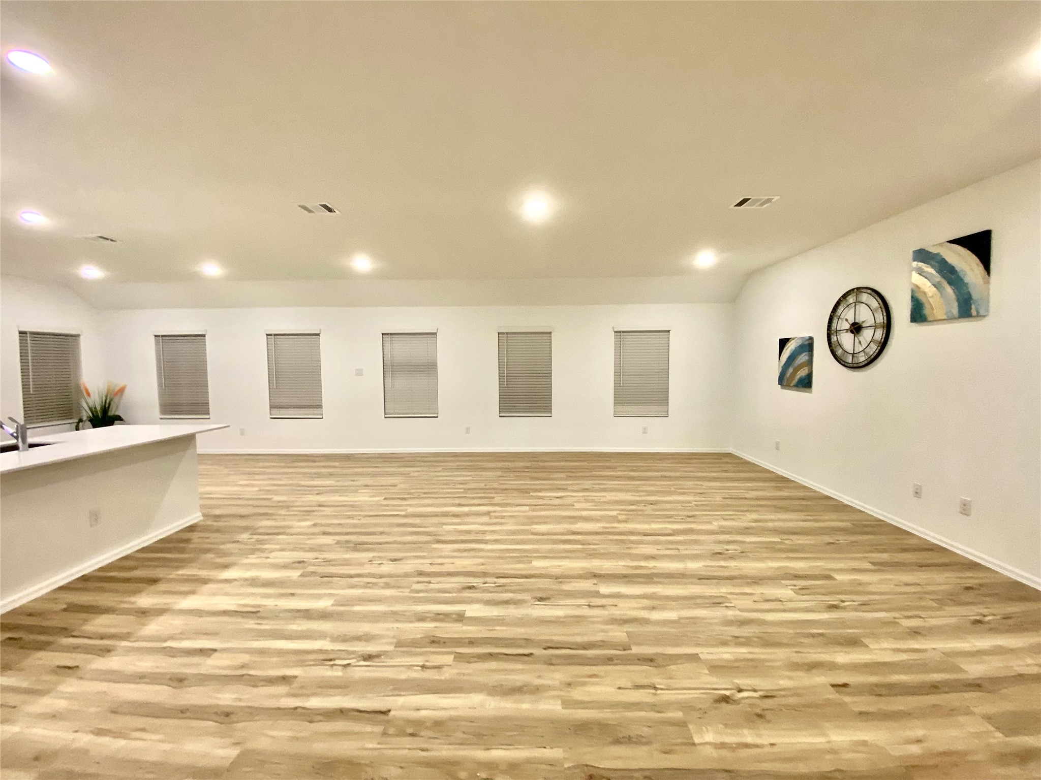 3724 Moss Hl Road Rosenberg, TX 77469 - Photo 9 of 29 Spacious open-plan area with light wood flooring, recessed lighting, and neutral walls. The space includes a modern kitchen counter on the left and windows with blinds. Decor features a large wall clock and abstract art. Ideal for flexible living and entertainment.