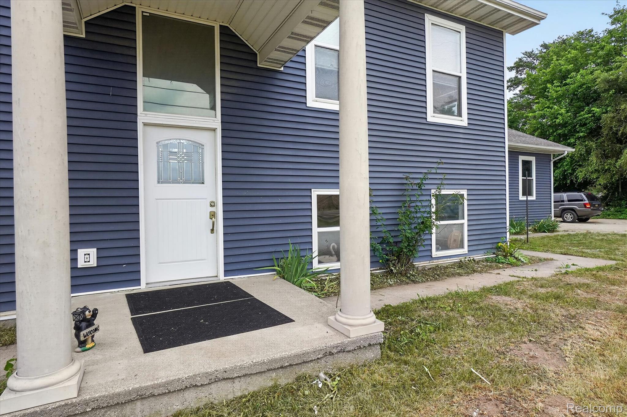 6524 Kelly Road Flushing, MI 48433 - Photo 6 of 47 Blue siding exterior featuring a white paneled entry door with decorative glass, an overhead transom window, and a concrete porch with substantial columns