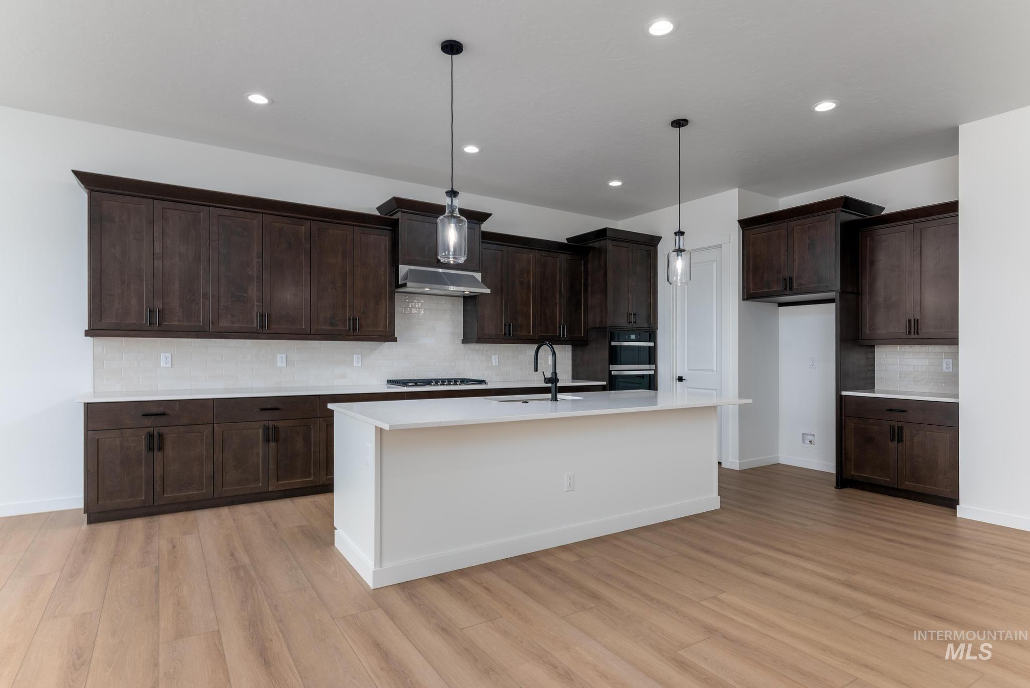8459 South Country Wind Avenue Meridian, ID 83642 - Photo 5 of 11 Kitchen with dark wood finish cabinetry, a center island with sink, pendant lighting, backsplash, and light wood-style flooring