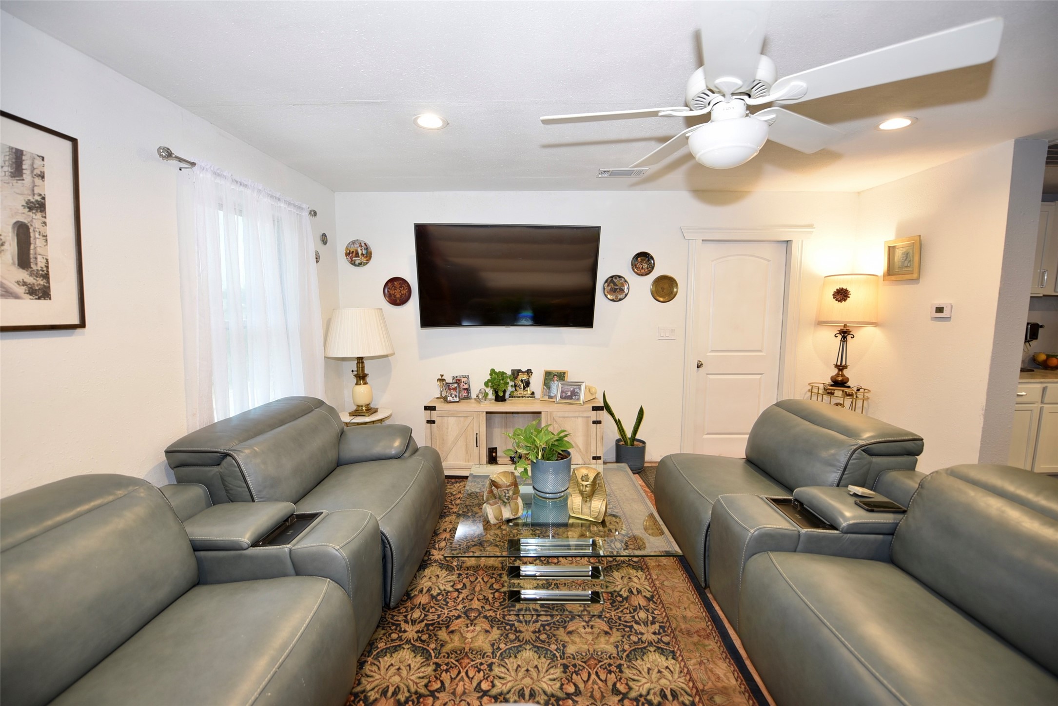 382 Road 5251 Cleveland, TX 77327 - Photo 5 of 22 Cozy living room with modern gray leather seating, a large wall-mounted TV, and ceiling fan. Features a glass coffee table, decorative plants, and soft lighting, creating a welcoming atmosphere.